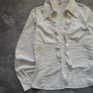 May include: White button-down shirt with long sleeves. The shirt features a collared neckline, decorative embroidery, and pleated and ruffled details. The shirt has a fitted waist and a button-up front.