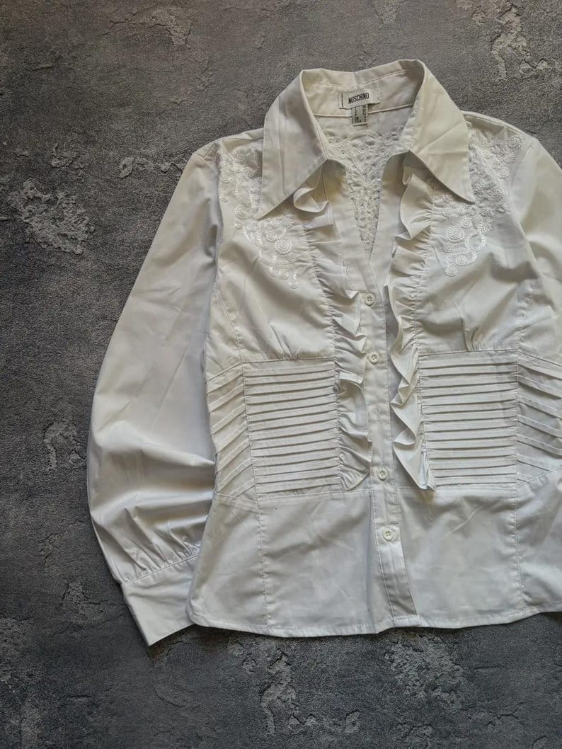 May include: White button-down shirt with long sleeves. The shirt features a collared neckline, decorative embroidery, and pleated and ruffled details. The shirt has a fitted waist and a button-up front.