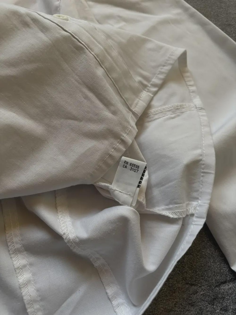 May include: Close-up of a white button-down shirt. The shirt is made of a smooth, crisp fabric. The tag reads RN 68596 CA 31127.