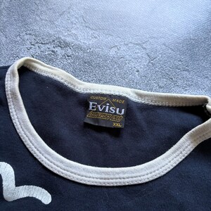 Puede incluir: Primer plano de una camiseta azul marino con ribete blanco en el cuello y las mangas. La etiqueta dice "CUSTOM MADE EVISU EVISU RANCH & CO XXL". La camiseta tiene un dise&ntilde;o gr&aacute;fico blanco en la parte delantera.