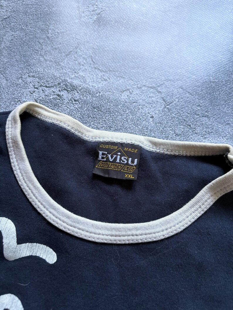 Puede incluir: Primer plano de una camiseta azul marino con ribete blanco en el cuello y las mangas. La etiqueta dice "CUSTOM MADE EVISU EVISU RANCH & CO XXL". La camiseta tiene un dise&ntilde;o gr&aacute;fico blanco en la parte delantera.
