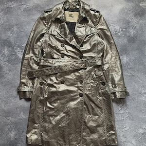 May include: A metallic silver double-breasted trench coat with a textured finish, featuring a wide collar, shoulder epaulets, a matching waist belt with a buckle, and buttoned cuffs. The interior label displays the Burberry brand name and logo.