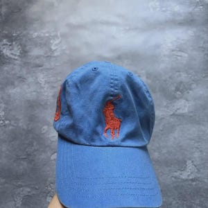 Peut inclure: Une casquette de baseball bleu clair avec un logo orange brodé représentant un joueur de polo sur un cheval sur le panneau avant. La casquette présente une visière incurvée avec des coutures visibles et une calotte structurée avec des œillets.