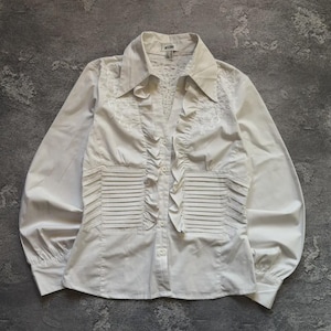 May include: White long-sleeve blouse with a collared neckline and button-down front. The blouse features decorative ruffles down the front and pleated details on the bodice. The sleeves are long and gathered at the cuffs. The blouse is made of a lightweight fabric.