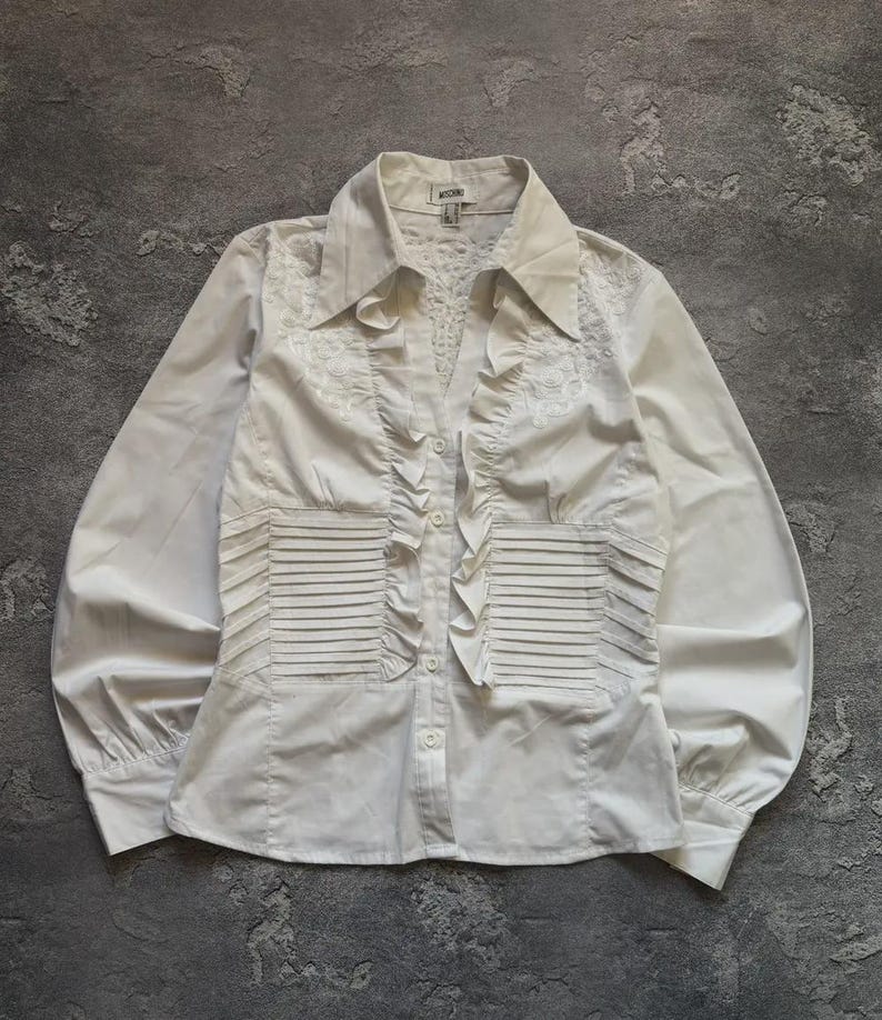 May include: White long-sleeve blouse with a collared neckline and button-down front. The blouse features decorative ruffles down the front and pleated details on the bodice. The sleeves are long and gathered at the cuffs. The blouse is made of a lightweight fabric.