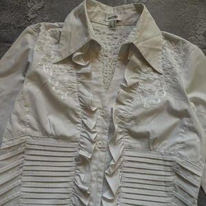 May include: A white button-down shirt with a collared neckline and long sleeves. The shirt features decorative embroidery on the upper chest and ruffled detailing down the front. The brand name "MOSCHINO" is visible on the label.