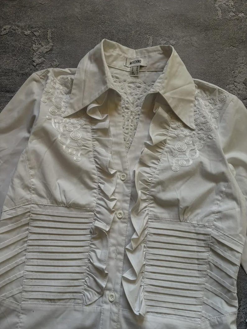 May include: A white button-down shirt with a collared neckline and long sleeves. The shirt features decorative embroidery on the upper chest and ruffled detailing down the front. The brand name "MOSCHINO" is visible on the label.