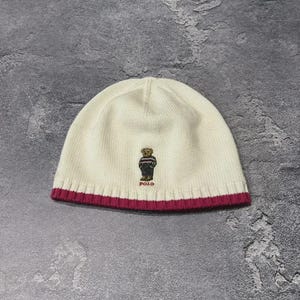 May include: A cream-colored knit beanie featuring a ribbed hem in a deep magenta hue. Centered on the front is an embroidered graphic of a teddy bear wearing a striped sweater and dark pants, with the word POLO stitched in red underneath the bear.