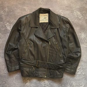 May include: A dark brown leather jacket with a zipper closure and a belt. The jacket has multiple pockets and a collar. The label reads "REAL LEATHER" and "joy AUTHENTICS".