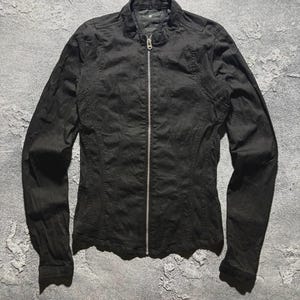 May include: A black jacket with a stand-up collar and a full-length silver zipper. The jacket has long sleeves and a tailored fit, with seams running vertically down the front and back. The fabric appears to be a lightweight material.