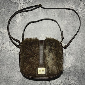 May include: A brown crossbody bag with a faux fur front panel. The bag has a leather strap and a rectangular clasp. The bag's dimensions are not visible in the image.