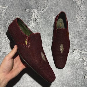 May include: A pair of burgundy slip-on shoes with a textured surface. Each shoe features a small, diamond-shaped embellishment on the vamp and a gold zipper detail. The shoes are displayed on a gray surface.