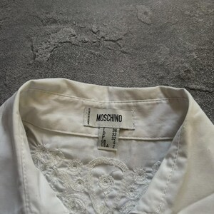 May include: Close-up of a white collared shirt with intricate floral embroidery on the front. The shirt features a label with the brand name "MOSCHINO" and size details. The shirt is made in Italy.