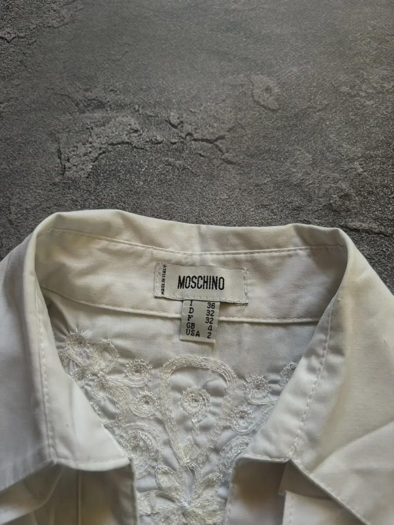 May include: Close-up of a white collared shirt with intricate floral embroidery on the front. The shirt features a label with the brand name "MOSCHINO" and size details. The shirt is made in Italy.