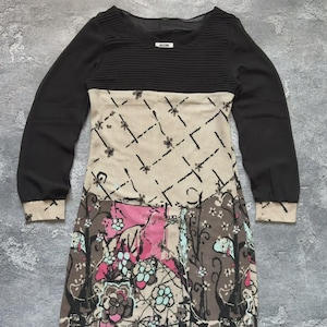 May include: A long-sleeved dress with a black bodice and sleeves. The dress features a beige midsection with a black geometric pattern and a floral print skirt in pink, brown, and blue. The dress has a round neckline and a label.