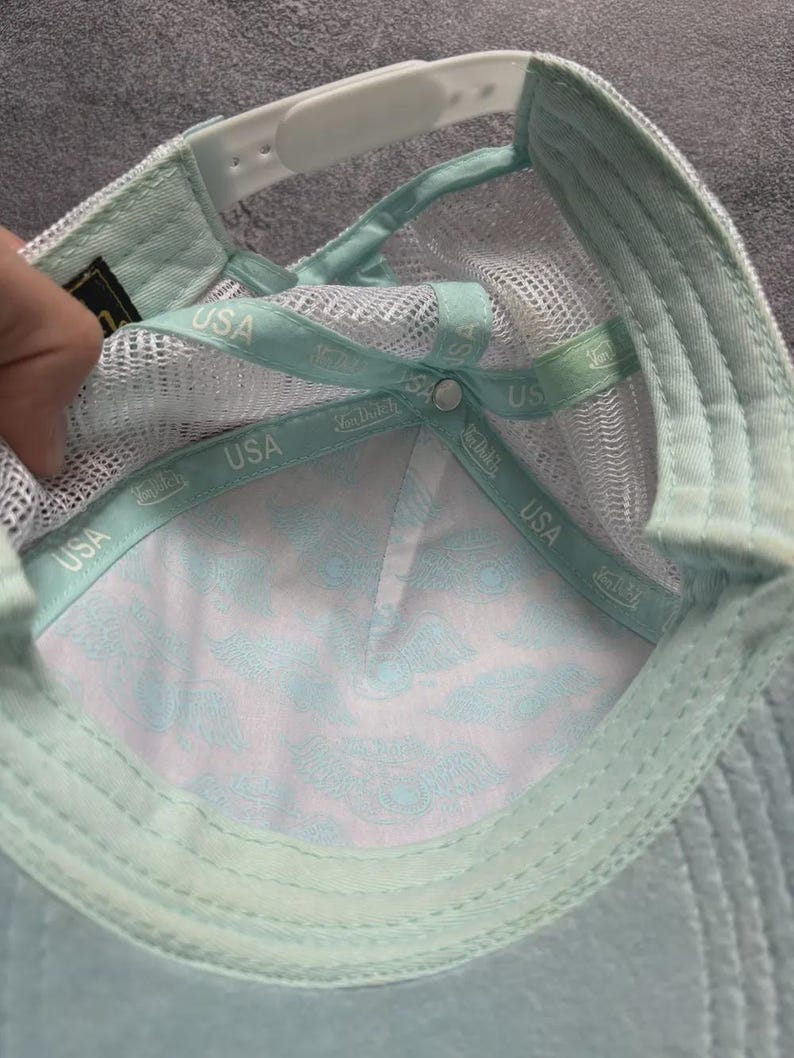 May include: A light blue and white trucker hat with a mesh back and a curved brim. The interior features a patterned design and a white adjustable strap. The hat has the text "USA" on the interior band.