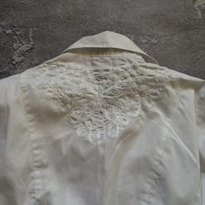 May include: A white collared shirt with intricate, embroidered detailing across the upper back. The embroidery is a delicate, floral-inspired design. The shirt is made of a smooth, possibly satin-like fabric.