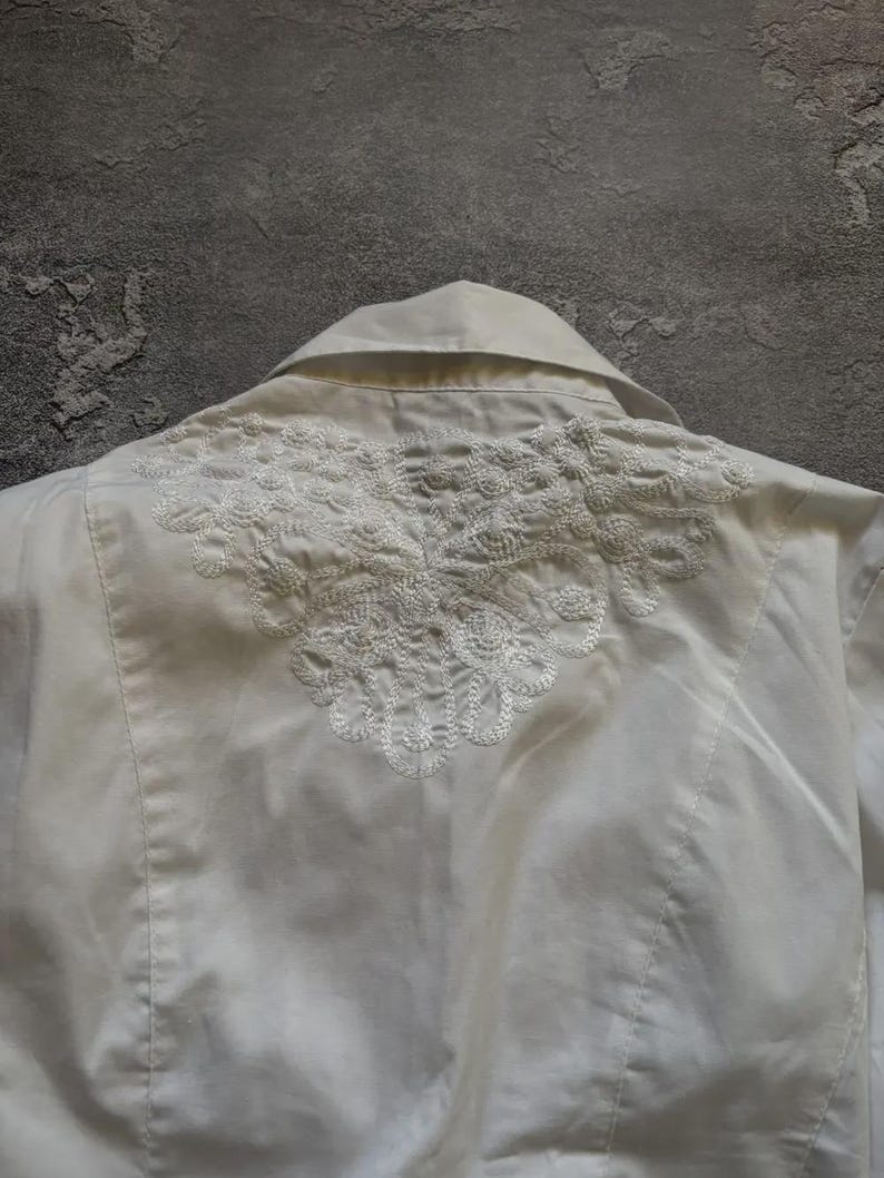 May include: A white collared shirt with intricate, embroidered detailing across the upper back. The embroidery is a delicate, floral-inspired design. The shirt is made of a smooth, possibly satin-like fabric.