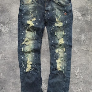 May include: Distressed blue denim jeans with ripped details and white paint splatter. The jeans have a button fly and a small logo patch near the front pocket. A red emblem is visible on the back pocket.