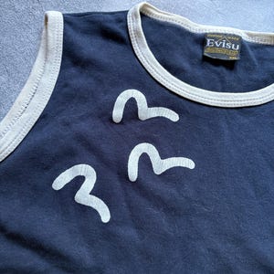 Puede incluir: Camiseta sin mangas azul marino con ribete crema y tres dise&ntilde;os blancos en forma de ola. La camiseta sin mangas tiene una etiqueta que dice "Evisu Custom Made". Es una camiseta sin mangas.