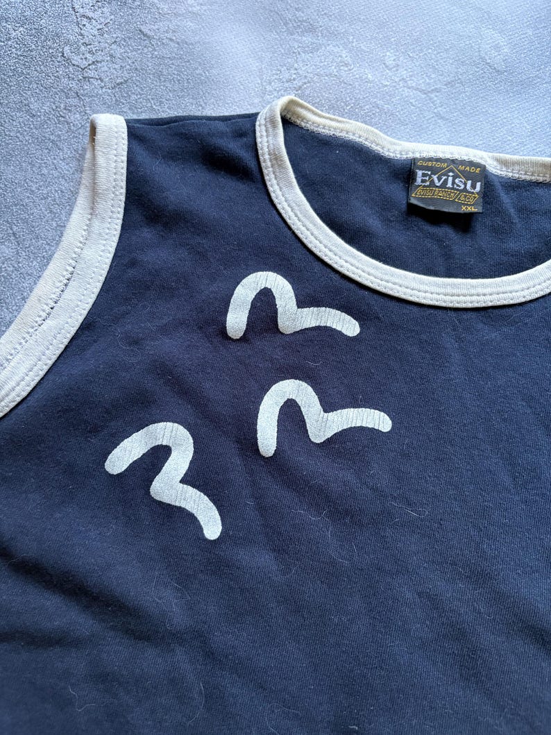 Puede incluir: Camiseta sin mangas azul marino con ribete crema y tres dise&ntilde;os blancos en forma de ola. La camiseta sin mangas tiene una etiqueta que dice "Evisu Custom Made". Es una camiseta sin mangas.
