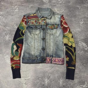 Puede incluir: Una chaqueta de mezclilla azul claro con mangas largas de punto sobre un fondo azul marino oscuro, decoradas con un gran patrón floral colorido. La chaqueta incluye cuello, cierre de botones, dos bolsillos en el pecho y bordados.