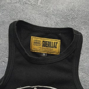 Może przedstawiać: Czarny podkoszulek z okrągłym dekoltem i wzorem o fakturze. Prostokątna etykieta z napisem "GUERILLAZ" i "CORTEZ" jest wszyta na dekolcie. Etykieta zawiera r&oacute;wnież tekst "The Blood of Battle, is Thicker than the Water of the Womb".