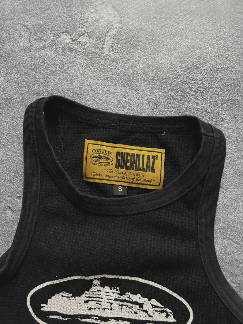 Może przedstawiać: Czarny podkoszulek z okrągłym dekoltem i wzorem o fakturze. Prostokątna etykieta z napisem "GUERILLAZ" i "CORTEZ" jest wszyta na dekolcie. Etykieta zawiera r&oacute;wnież tekst "The Blood of Battle, is Thicker than the Water of the Womb".