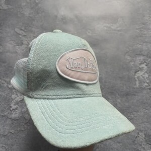 May include: A light blue trucker hat with a curved brim and mesh back. The hat features a patch with the text "Von Dutch" in a cream-colored oval.