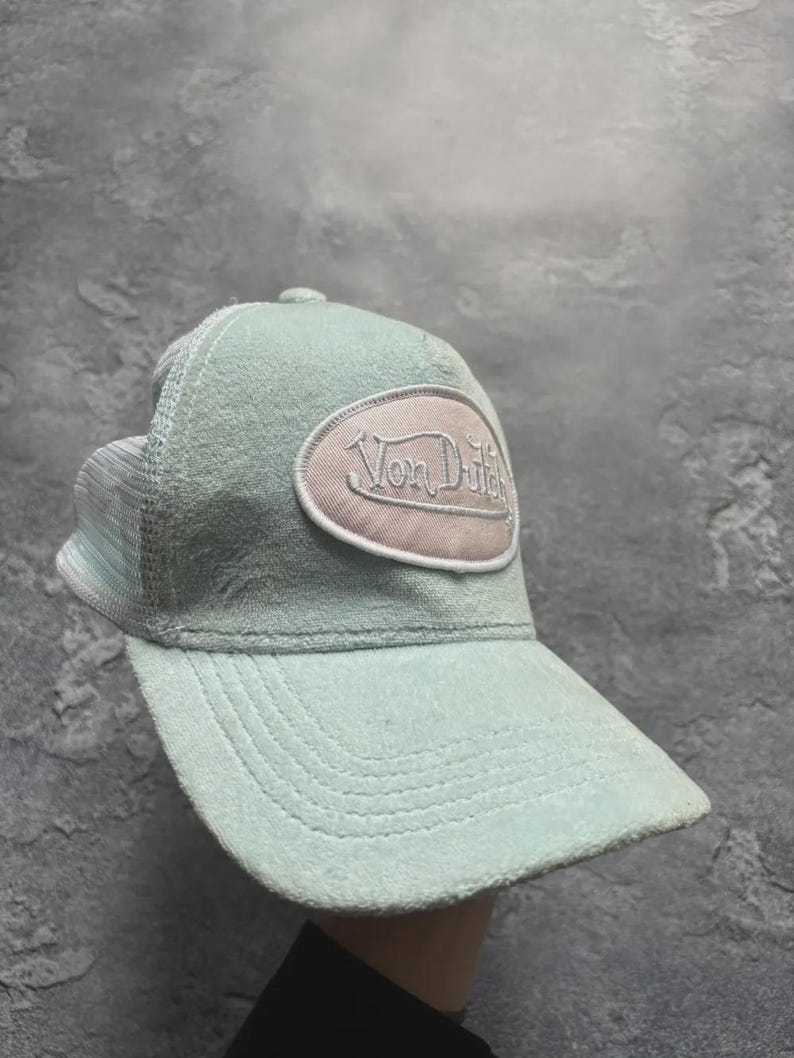 May include: A light blue trucker hat with a curved brim and mesh back. The hat features a patch with the text "Von Dutch" in a cream-colored oval.