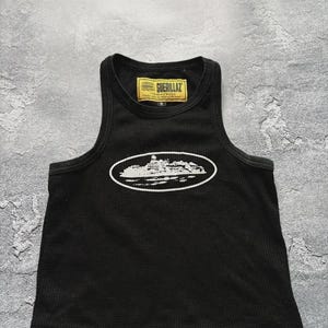 Może przedstawiać: Czarny tank top z fakturą wafelkową. Top posiada białą owalną grafikę z projektem budynku oraz napis "GUERILLAZ" na ż&oacute;łtej etykiecie.