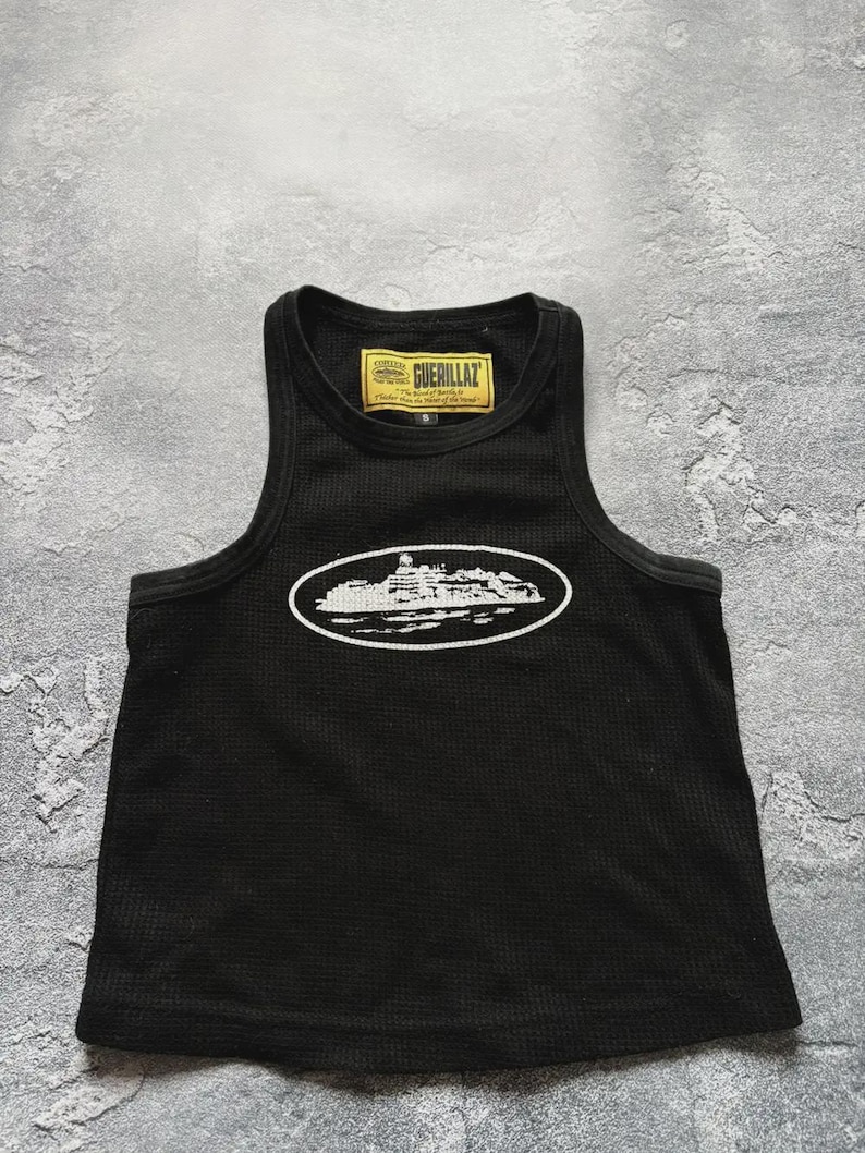 Może przedstawiać: Czarny tank top z fakturą wafelkową. Top posiada białą owalną grafikę z projektem budynku oraz napis "GUERILLAZ" na ż&oacute;łtej etykiecie.