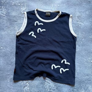 Puede incluir: Camiseta sin mangas azul marino con ribete blanco y gr&aacute;ficos de p&aacute;jaros blancos. La camiseta tiene la marca "Evisu" en el cuello. Una camiseta informal para el uso diario.