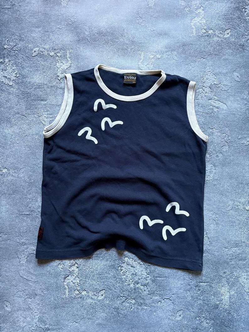 Puede incluir: Camiseta sin mangas azul marino con ribete blanco y gr&aacute;ficos de p&aacute;jaros blancos. La camiseta tiene la marca "Evisu" en el cuello. Una camiseta informal para el uso diario.