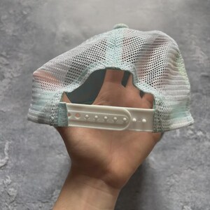 May include: A light blue trucker hat with a white mesh back and a white adjustable strap. The hat has a curved brim and a light blue front panel. The hat is designed to be worn with the brim facing forward.