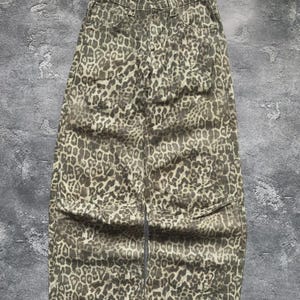 Puede incluir: Vaqueros de talle alto y pernera ancha con estampado de leopardo en tonos marrón y beige. Los pantalones tienen un diseño clásico de cinco bolsillos y cierre de botón y cremallera. La tela parece ser un denim resistente.