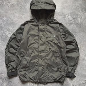 May include: An olive green hooded jacket featuring a front zipper closure with a snap-button storm flap, two slanted zippered pockets, and adjustable cuffs. The Haglöfs brand logo is embroidered in white on the upper left chest area.