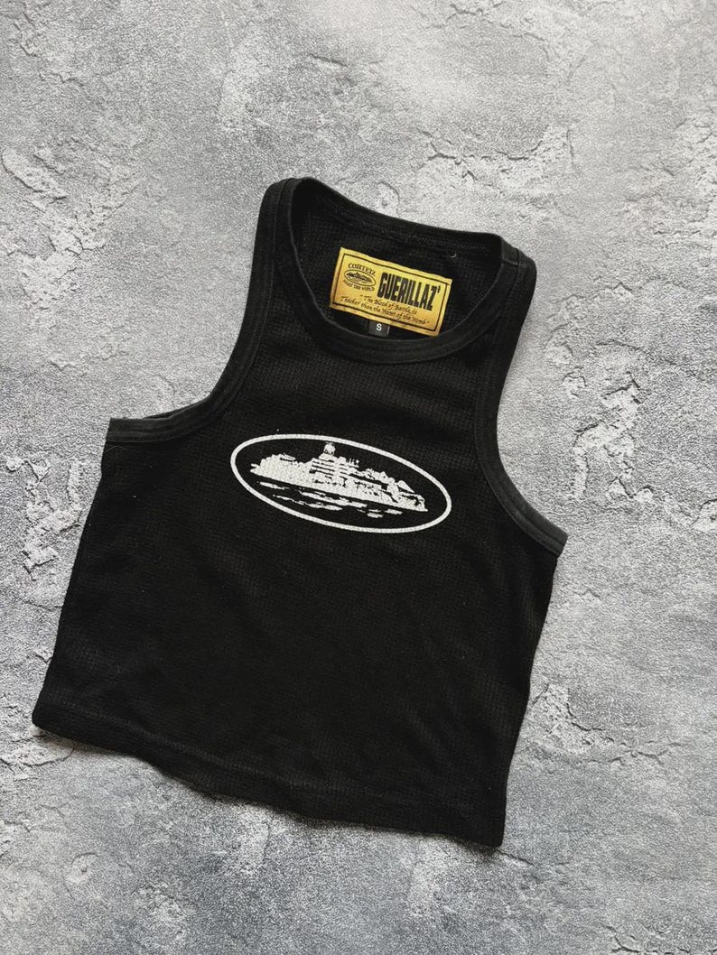 Może przedstawiać: Czarny top z białym owalnym nadrukiem przedstawiającym zarys budynku. Top ma prążkowaną fakturę i małą ż&oacute;łtą metkę z napisem "GUERILLAZ". Top jest w rozmiarze S.