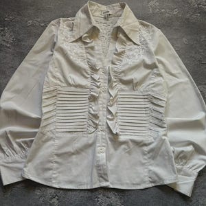 May include: A white button-down shirt with long sleeves. The shirt features a collared neckline, decorative ruffles down the front, and pleated details on the front. The fabric appears to be a smooth, possibly satin-like material.