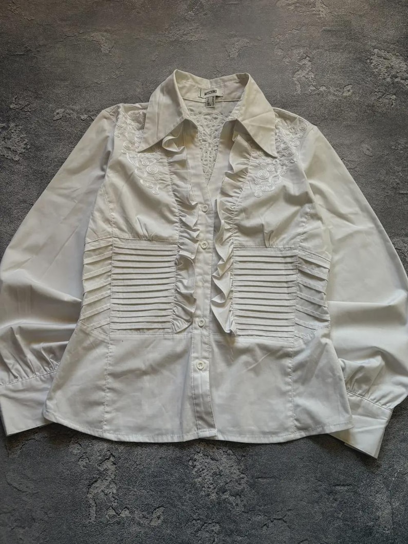 May include: A white button-down shirt with long sleeves. The shirt features a collared neckline, decorative ruffles down the front, and pleated details on the front. The fabric appears to be a smooth, possibly satin-like material.