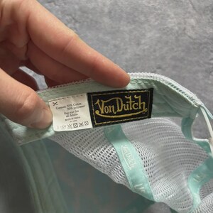 May include: A Von Dutch baseball cap in light blue and white. The cap features a mesh back and a curved brim. The Von Dutch logo is displayed on a black and gold patch. The cap is made of cotton and polyester.