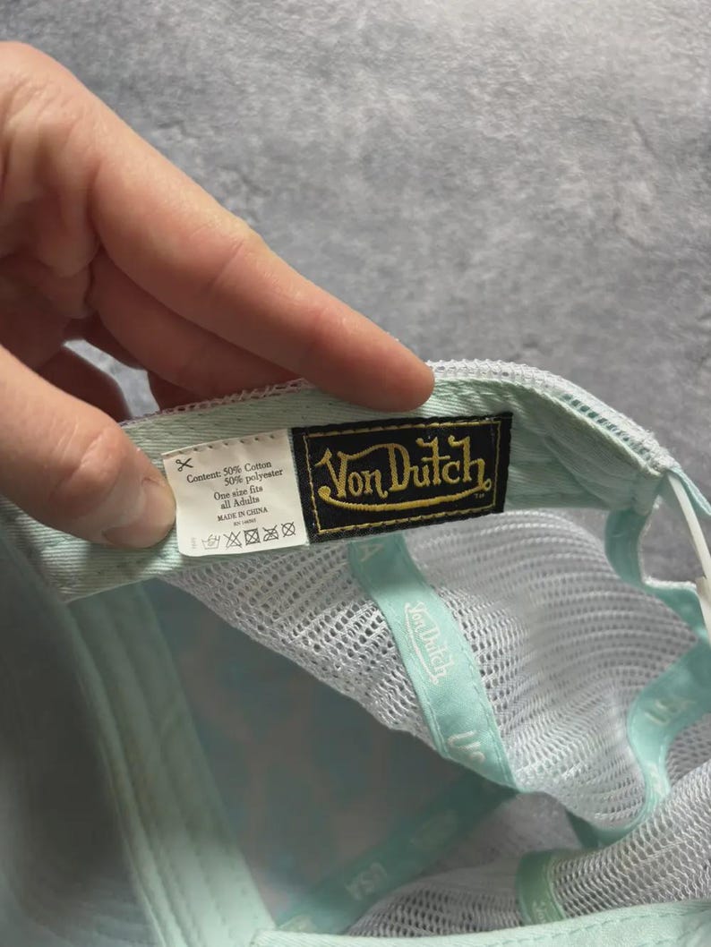 May include: A Von Dutch baseball cap in light blue and white. The cap features a mesh back and a curved brim. The Von Dutch logo is displayed on a black and gold patch. The cap is made of cotton and polyester.
