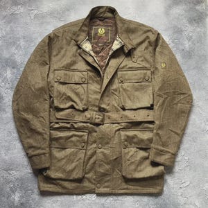 May include: A brown, long-sleeved, belted jacket featuring four large front pockets with snap closures, a high collar, and a quilted brown interior lining. The brand label Belstaff is visible on the inner neck and a small logo patch on the sleeve.