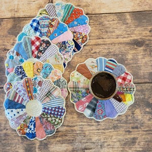 May include: A collection of handmade, flower-shaped coasters. Each coaster is crafted from a patchwork of colorful fabric scraps, featuring various patterns and textures. One coaster holds a cup of coffee. The coasters are arranged on a wooden surface.