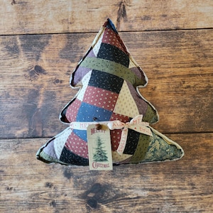 May include: A stuffed fabric Christmas tree ornament featuring a patchwork design of various patterned fabrics in shades of red, blue, green, and cream. A cream ribbon tied around the base holds a tag with a tree illustration and the text Merry Christmas.