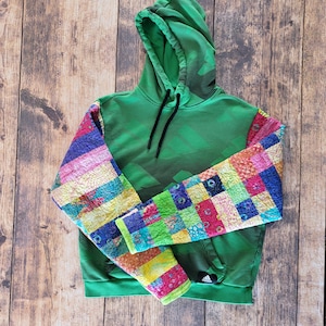 May include: Green hooded sweatshirt with black Adidas logo and accents. The sleeves are patchwork with a variety of colorful squares. The drawstring is black.