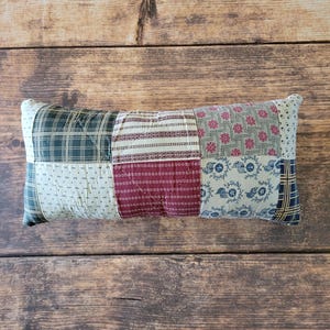 May include: A rectangular patchwork pillow featuring a variety of fabric patterns including dark green plaid, cream with small black marks, burgundy stripes, floral motifs in pink and blue, and a navy blue plaid section, all stitched together with quilting.