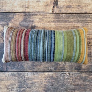 May include: A rectangular throw pillow featuring a vertical striped pattern in shades of brown, maroon, blue, green, and yellow. The fabric has a quilted texture with visible stitching lines running parallel to the stripes across the entire surface.