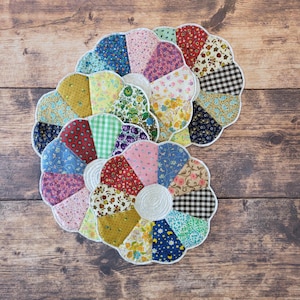 May include: A set of two flower-shaped fabric coasters or trivets, each constructed from multiple wedge-shaped segments of various patterned cotton fabrics including floral, gingham, and geometric prints, with a white center and white trim.