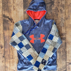 May include: Blue hoodie with a coral-colored interior and a coral logo. The sleeves feature a patchwork design with squares of blue, white, and beige fabric. The hoodie has a drawstring and a front pocket.